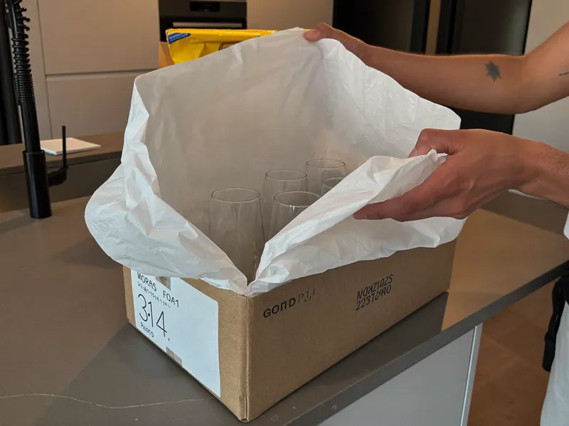 Packer wrapping glassware in white paper and placing it in a carton on a kitchen counter